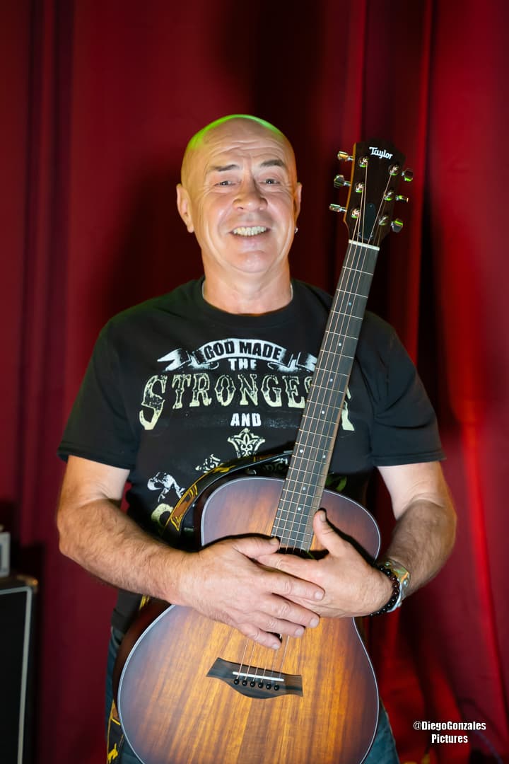 Oleg Sweet smiling with his acoustic guitar against a red stage curtain